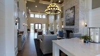 This single-story space makes an immediate impression with its dramatically tall ceilings framed by coffered detailing and clerestory windows that flood the interior with natural light. The grand scale of the room is anchored by two statement chandeliers — a large, tiered candle-style fixture and a smaller crystal chandelier — that add warmth and visual drama overhead. Dark hardwood flooring contrasts beautifully with the light, neutral wall tones, creating a refined yet inviting atmosphere. A floor-to-ceiling stacked stone fireplace serves as a striking focal point along one wall, complemented by a large landscape painting that enhances the room's artistic character. The seating area features patterned and upholstered armchairs arranged around a central coffee table, encouraging comfortable conversation. Arched French doors at the far end draw the eye outward and invite natural light deep into the space. Wainscoting and architectural millwork throughout add classic elegance, while a white countertop surface in the foreground suggests a functional service or reception area. Overall, the space blends traditional architecture with sophisticated interior design.