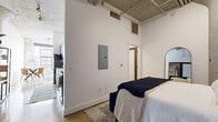 Located in the heart of downtown Los Angeles, this 1-bedroom, 1-bathroom loft at 217 E 8th St blends industrial character with modern comfort. The space is defined by its soaring exposed concrete ceilings with visible ductwork and pipes, original concrete columns, and polished concrete and tile flooring — hallmarks of classic LA adaptive reuse architecture.

The open living area offers a generous, light-filled layout with tall ceilings that amplify the sense of space, complemented by clean white walls and a warm, minimalist aesthetic. The kitchen is well-appointed with dark wood cabinetry, white quartz countertops, a marble-style tile backsplash, and stainless steel appliances including a dishwasher, range, and microwave.

The bedroom is a comfortable retreat featuring an arched concrete column accent, a sliding glass-panel closet, and the same soaring ceiling height found throughout. The bathroom is clean and modern, finished with large-format floor tiles, a sleek vanity, and a tub-shower combination enclosed by glass and clad in dark mosaic tile. A built-in electrical panel is discreetly tucked into the wall. This loft offers a distinctive urban living experience in one of LA's most vibrant downtown neighborhoods.
