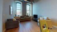 This single-floor residence showcases a compelling blend of industrial character and modern comfort. The living area commands immediate attention with its dramatically tall arched windows, allowing abundant natural light to flood the space and highlighting the warm hardwood flooring that runs throughout. Exposed structural elements, including a prominent wooden post, add a distinctive loft-like aesthetic to the open-concept layout.

The kitchen is well-appointed with warm maple cabinetry, granite countertops, a full suite of stainless steel appliances — including a range, over-the-range microwave, and refrigerator — and a peninsula that serves as both a prep area and casual dining spot with bar seating. The entry hallway connects the main living areas to the private quarters with ease.

The bedroom continues the home's design language with wainscoting along the lower walls, large gridded windows that bring in generous daylight, a ceiling fan, and the same hardwood flooring found throughout. The bathroom features warm wood cabinetry, a granite-topped vanity with a large mirror, and hardwood-style flooring, maintaining a cohesive and polished finish. Overall, the space offers a bright, airy ambiance with architectural details that give it a unique and distinctive character.