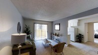 This single-story residence features a bright and functional open-concept layout. The living room serves as the central gathering space, finished with neutral carpet flooring and light gray walls that create a calm, understated atmosphere. A textured popcorn ceiling adds a classic touch overhead. The room is furnished with a comfortable sofa accented by decorative pillows, a wicker accent chair, and a metal-framed coffee table, together creating a relaxed yet coordinated aesthetic. A decorative wall mirror and console table with tasteful accents add character to the space.

A sliding glass door fills the living area with natural light and opens onto a private balcony, offering views of the surrounding exterior. This connection to the outdoors enhances the sense of openness within the space. Adjacent to the living room, the kitchen is partially visible through an open pass-through, showcasing white cabinetry, white appliances including a refrigerator, and light countertops. The seamless flow between the living and kitchen areas supports an efficient, easy-to-navigate floor plan. Overall, the space offers a practical and comfortable environment with a clean, modest design.