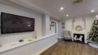This thoughtfully designed single-floor space combines classic and contemporary elements to create a refined, inviting environment. The main living area showcases rich, dark hardwood-style flooring that runs throughout, providing a striking contrast against the crisp white walls and bright trim work. Crown molding adds an elegant architectural detail along the ceiling, lending a polished, upscale character to the space.

A built-in media wall with a large wall-mounted television serves as a central focal point, complemented by a sleek, low-profile white media console below. Recessed lighting in the ceiling ensures even, ambient illumination throughout. On the opposite end, a classic white-painted brick fireplace anchors the room with warmth and character, adorned with decorative accents and framed artwork arranged neatly on the surrounding wall.

A cozy accent chair with a patterned upholstery sits adjacent to the fireplace, creating an intimate reading or conversation nook. Natural light filters in through window treatments, brightening the space without sacrificing privacy. A hallway visible in the background hints at additional rooms beyond, suggesting a well-organized and functional layout. Overall, the space exudes a clean, welcoming aesthetic that balances comfort with tasteful design.