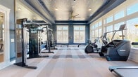 Located at 900 Mountaintop Ave in Gainesville, GA, this dedicated exercise room delivers a premium fitness experience within a thoughtfully designed single-level space. The room's most striking architectural feature is its ornate tray ceiling, adorned with a detailed decorative pattern and complemented by a stylish gold ceiling fan and recessed lighting that together create an atmosphere well above the ordinary gym setting. Deep navy blue walls add a bold, energizing character throughout, while full-length mirrors along one wall visually expand the space and support form-focused training. Large, multi-pane windows line two walls, flooding the room with abundant natural light and offering views of the surrounding wooded landscape and outdoor pergola structure. The floor is finished with a custom patterned carpet tile in soft blue, gray, and cream tones, providing both comfort and a distinctive aesthetic. The exercise room is well-equipped with a diverse array of fitness equipment, including treadmills, an elliptical trainer, a stationary bike, a rowing machine, a multi-station cable machine, a weight rack, and a flat bench — offering comprehensive cardio and strength training options. The combination of refined design details, ample natural light, and a full range of equipment makes this fitness space both functional and visually impressive.