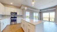 This single-floor space is designed with a clean, contemporary aesthetic that emphasizes functionality and natural light. The open-concept kitchen serves as the centerpiece, showcasing white shaker-style cabinetry with brushed gold hardware, a generous island with a white quartz countertop, and a full suite of stainless steel appliances including a range, built-in microwave, and refrigerator. Warm-toned hardwood-style flooring runs throughout the main living areas, complementing the neutral gray wall tones. Pendant lighting with glass fixtures hangs above the island, adding a refined touch to the space. Large windows and sliding glass doors frame impressive views of lush treetops and a waterway beyond, flooding the kitchen and adjoining living area with abundant natural light. The bathrooms are finished with white shaker vanities, quartz countertops, and hardwood-style flooring, maintaining a consistent modern palette. One bathroom features a tub-shower combination, while another offers additional tall cabinetry for storage. The bedrooms are carpeted for comfort and include expansive windows that capture scenic outdoor views, creating bright and inviting retreats throughout the home.