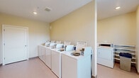 This shared laundry facility is a compact, single-floor space designed for efficient, communal use. The room is laid out to maximize functionality, with a row of top-loading washing machines aligned along one wall and a set of dryers positioned nearby, providing ample capacity for multiple users. The machines are uniformly white, contributing to the room's clean, orderly appearance.

The walls are finished in a soft, warm yellow tone, lending a bright and welcoming atmosphere to what is typically a utilitarian space. Light-toned vinyl flooring runs throughout, offering a practical and easy-to-maintain surface. Recessed lighting and ceiling-mounted ventilation fixtures ensure the room remains well-lit and properly ventilated during use.

A large window fitted with horizontal blinds allows natural light to fill the space, offering views of the outdoor surroundings and enhancing the room's overall brightness. A solid entry door, equipped with a keypad lock, provides secure, controlled access to the facility. Structural columns are integrated into the layout without significantly impeding the usable floor area.

Overall, the space balances practicality with a tidy, well-maintained aesthetic, making it a functional and accessible laundry solution for shared use.