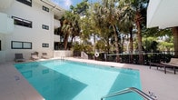 This single-floor accessible space is part of a modern, white-exterior multi-story building nestled within a lush tropical landscape. The standout feature of the property is the generously sized outdoor swimming pool, which boasts crystal-clear turquoise water and is surrounded by a clean, light-tiled pool deck furnished with lounge chairs for relaxation. The pool area is enclosed by a black metal safety fence and is framed by towering palm trees and dense mature foliage, creating a serene, resort-like atmosphere. Safety amenities such as a life ring are visibly present poolside.

The building's exterior showcases a clean, contemporary white finish with large sliding and fixed black-framed windows that allow natural light into the interior spaces. The open-air corridors are finished with large-format light stone tiles and feature sturdy black metal railings, providing both safety and an aesthetic complement to the building's modern design. Access between levels is facilitated by well-lit stairwells. Interior utility areas present bright, open white-walled rooms with light wood-style flooring and sliding glass doors overlooking the outdoor amenities. The overall ambiance is bright, clean, and inviting, with a strong emphasis on indoor-outdoor connectivity enhanced by the surrounding natural greenery.