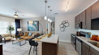 Located at 13555 Yellow Fern Way in Riverview, Hillsborough County, Florida, this single-story residence showcases a thoughtfully designed open-concept layout with modern finishes throughout. The kitchen serves as a central highlight, featuring dark wood cabinetry, granite countertops, a subway tile backsplash, stainless steel appliances, and a large island with pendant lighting overhead — ideal for both meal preparation and casual dining. Light wood-style flooring extends through the main living areas, complementing the neutral gray walls and creating a bright, cohesive aesthetic. The living room offers generous natural light through large windows dressed with layered curtains and plantation shutters, a ceiling fan, and ample space for comfortable seating arrangements. The primary bedroom provides a warm, inviting retreat with a richly toned accent wall, plush carpeting, and room for a king-sized bed. A second bedroom also features large windows and neutral finishes. Both bathrooms are appointed with dark wood vanities, granite countertops, framed mirrors, and tile flooring. The primary bath includes a tub and shower combination with stone-look tile surround, while the secondary bath offers a refined vanity with generous storage. The space as a whole presents a clean, contemporary style with upscale detailing throughout.