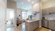 This single-floor residence in Savannah, GA, showcases a contemporary open-concept design with clean lines and modern finishes throughout. The entryway features a sleek dark wood door with a keypad lock, opening into a space defined by warm wood-look flooring and neutral tones.

The kitchen serves as a central focal point, anchored by a granite-topped island ideal for dining or meal prep. Taupe wood cabinetry, subway tile backsplash, stainless steel appliances—including a French door refrigerator, range, microwave, and dishwasher—and track lighting combine for a highly functional and stylish cooking space. Open metal shelving adds an industrial touch.

The living area flows seamlessly from the kitchen, featuring notably tall ceilings that amplify the sense of space. Large French doors flood the room with natural light and provide access to a private balcony. The bedroom offers plush carpeting, multiple windows with ample natural light, and sufficient space for a full furniture arrangement including a desk area. Throughout the residence, the cohesive neutral palette, elevated ceilings, and thoughtful layout create a bright, comfortable, and modern living environment.
