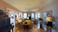 This charming single-floor residence exudes a timeless, classic character that is immediately evident upon entering the main living area. The living room serves as the heart of the space, showcasing rich hardwood floors that run throughout and lend a sense of warmth and continuity. Crown molding frames the ceiling, which features subtle textural detailing that adds a touch of traditional elegance to the room.

Multiple double-hung windows line the walls, flooding the interior with generous natural light and offering pleasant views of the surrounding greenery outdoors. The soft, warm tones of the walls complement the inviting arrangement of plush, earth-toned sofas and furnishings, creating a cozy yet refined atmosphere. A classic fireplace mantel with an ornate mirror serves as a focal point, flanked by table lamps that cast a gentle ambient glow throughout the space.

A writing desk and chair are thoughtfully positioned within the living area, adding both function and charm. Decorative accents, including framed artwork, fresh floral arrangements, and personal mementos, contribute to the lived-in, homey character of the space. An open doorway connects to an adjacent room, hinting at further well-appointed living areas beyond. Overall, this space blends traditional architectural details with comfortable, curated furnishings.
