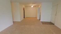 This single-floor space features an open-concept living area finished with wall-to-wall neutral-toned carpeting that lends a clean, understated aesthetic throughout. The main living area is generously proportioned, offering ample room for a variety of furniture arrangements. A ceiling fan with a built-in light fixture is centrally mounted overhead, providing both air circulation and ambient lighting to the space.

The layout connects seamlessly to additional areas of the unit. An opening along one wall leads to what appears to be a kitchen, where a stainless steel refrigerator is visible, suggesting a functional and equipped cooking space. A set of bifold doors nearby likely conceals a closet, offering convenient storage within easy reach of the main living area. Adjacent to this, a transition to hard surface flooring hints at a separate room or entryway, adding practical variety to the overall floor plan.

The walls are painted in a soft, neutral tone throughout, complementing the light carpeting and contributing to a bright, cohesive feel across the space. The straightforward, open design makes this a versatile and adaptable living environment.