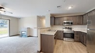 This single-floor residence offers a thoughtfully designed open-concept layout that seamlessly connects the kitchen and living areas. The kitchen is a functional highlight, featuring rich dark taupe cabinetry, light granite-style countertops, and a full suite of stainless steel appliances including a refrigerator, range, and over-the-range microwave. A peninsula with a double sink serves as a natural divider between the kitchen and the adjoining living space, which is finished with warm light wood-style flooring. The living room is anchored by a sleek tile-surround fireplace and ceiling fan, creating a cozy yet modern atmosphere. Built-in open shelving adds a decorative architectural touch near the transition between rooms. The bedrooms are generously sized, each equipped with ceiling fans and large windows that invite natural light and outdoor views. Plush wall-to-wall carpeting runs throughout the bedrooms, adding warmth and comfort underfoot. The bathroom is finished with matching taupe cabinetry, a wide vanity mirror, and granite-style countertops, maintaining a cohesive design palette throughout. A sliding glass door provides direct access to the outdoors, connecting the interior to surrounding green space.