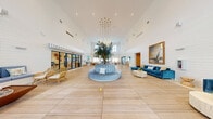 Located at 5475 N University in Lauderhill, Florida, this single-floor communal space offers an expansive, open-plan layout that exudes a refined coastal aesthetic. The grand lobby commands immediate attention with its dramatically soaring ceilings, which are accentuated by large clerestory windows that flood the interior with abundant natural light. At the heart of the space stands a striking live indoor tree encircled by a curved, channel-tufted blue sectional sofa, serving as a captivating focal point beneath a sculptural gold cluster chandelier. Light-toned wood-look tile flooring extends throughout, enhancing the bright, airy atmosphere. Multiple seating areas are thoughtfully arranged across the open floor, including plush navy blue velvet sofas, natural rattan accent chairs, and organic coffee tables, all unified by a consistent coastal palette of blues, whites, and warm naturals. Nautical-themed artwork, including sailboat paintings and ocean photography, adorns the shiplap-paneled walls. A separate lounge nook offers an intimate retreat with floor-to-ceiling sheer drapery and coastal-inspired furnishings. A distinct service area enclosed by glass doors beneath a striped awning adds functional depth to the space, completing this thoughtfully designed, resort-style environment.