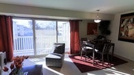 This single-floor residence offers a welcoming and open layout that seamlessly connects the living and dining areas. The space is flooded with natural light, courtesy of a large sliding glass door that spans much of the exterior wall, offering views of the surrounding outdoor scenery and providing direct access to a private deck. The living area is carpeted and provides a comfortable, relaxed setting, complemented by a patterned accent chair and warm, neutral wall tones. Rich burgundy drapery panels frame the sliding door, adding a bold contrast to the light-filled room. The dining area is situated adjacent to the living space and features a dark wood dining table paired with matching chairs, set atop a deep red area rug that anchors the space. A pendant light fixture hangs above the dining area, providing focused illumination and a touch of refined style. Decorative wall art and a potted plant add personality and warmth to the corner of the room. Together, the open connection between living and dining spaces, the abundance of natural light, and the cohesive design elements create a functional and inviting atmosphere throughout this single-floor home.