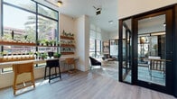 Located at 2600 N Orange Ave in Orlando, Florida, this contemporary single-floor commercial space combines modern design with functional versatility. Floor-to-ceiling windows flood the interior with natural light and offer views of the surrounding urban landscape, including lush palm trees, creating a bright and welcoming atmosphere throughout.

The open layout features light wood-look flooring in key areas and complementary textured carpet in lounge zones, tying together the space's cohesive aesthetic. A dedicated lounge area provides comfortable seating, accented by curated artwork — including a striking large-format black-and-white photography piece — and natural decorative elements such as vases with greenery.

A built-in kitchenette station includes open shelving stocked with books and décor, a coffee machine, a compact under-counter refrigerator, and a white countertop workspace, making it both practical and visually appealing. An adjacent window-side bar counter with stools and tiered wooden shelves lined with potted plants adds a vibrant, biophilic touch.

Sleek black-framed glass partitions and doors divide the space into distinct private areas, including enclosed offices visible with conference seating and display areas. Built-in shelving niches in the corridors add functional storage while maintaining the refined aesthetic. The overall ambiance is polished, professional, and thoughtfully designed for a modern work environment.
