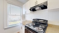 This two-floor space offers a well-organized layout spread across a main level and an upper level, making efficient use of every square foot. The main floor includes a kitchen, hallway, and bathroom, while the upper floor provides additional living area accessible via an interior staircase.

The kitchen is a standout feature, finished with crisp white cabinetry equipped with sleek bar-pull hardware and stone-look countertops. A black electric range with a stainless steel range hood serves as the focal point, complemented by a black dishwasher. Large windows with horizontal blinds allow generous natural light to fill the space, offering views of the surrounding greenery. The kitchen flooring is warm hardwood-style planking, adding a welcoming tone.

The bathroom continues the clean aesthetic with white walls, hardwood-style flooring, a white vanity, toilet, and a tiled shower-tub combination. A bright window keeps the room feeling open and airy, while a grab bar adds a practical accessibility element.

The upper level features plush carpeting and a sloped ceiling that gives the space a cozy, loft-like character. Recessed lighting and a small window provide illumination, while a half-wall railing overlooks the lower level, creating a sense of openness and connectivity between floors. Warm wood-trimmed windows on the main level rooms further enhance the space's inviting character.