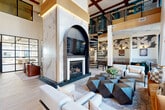 Located at 2500 Vantage Dr in Woodbridge, Virginia, this sophisticated communal lounge offers a generously scaled, single-floor layout designed to impress. The centerpiece of the space is a dramatic floor-to-ceiling marble fireplace surround, framed by a bold black arch that houses an embedded flat-screen display — a striking architectural focal point that commands attention from every seating area. Soaring double-height ceilings amplify the sense of grandeur, complemented by warm wood beam accents and ambient cove lighting that add depth and dimension overhead. A sculptural spiral staircase with sleek metal railings rises to an upper-level mezzanine, adding vertical drama and connectivity to the space. Light wide-plank flooring runs throughout, providing a clean, contemporary foundation. Multiple seating vignettes are thoughtfully arranged throughout — from plush lounge sofas and leather accent chairs to intimate booth-style dining nooks with marble-topped tables. Large floor-to-ceiling windows flood the space with natural light and offer open views of the surrounding community courtyard. Decorative wall art, bold textiles, and curated furnishings in rich tones of teal, cognac, and gold complete the polished, design-forward aesthetic of this inviting communal retreat.