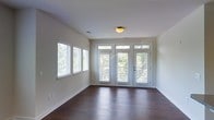 This single-floor space offers a clean, well-maintained interior defined by a thoughtful balance of natural light and classic finishes. The main living area is grounded by rich, dark hardwood floors that extend throughout, adding warmth and visual depth to the space. Smooth, neutral-toned walls provide a versatile backdrop that complements a variety of interior styles. A striking wall of windows, complete with white horizontal blinds, lines one side of the room, while a set of glass-paneled French doors with transom windows above allows generous views of the lush, tree-lined surroundings outside. The abundance of glazing ensures the space is consistently bathed in natural light throughout the day. A flush-mount ceiling light fixture provides additional illumination. Subtle architectural details — including clean baseboards, visible HVAC vents, and crisp white window trim — contribute to a polished, move-in-ready feel. The open layout creates a sense of spaciousness, offering flexibility for various furniture arrangements. Overall, the space presents a bright, inviting atmosphere with durable finishes and a strong connection to the natural environment just beyond its walls.