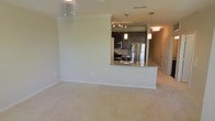 This single-floor space showcases an open-concept layout that seamlessly connects the living area to the kitchen. The living area is finished with plush neutral-toned carpet and features crown molding along the ceiling, adding a polished architectural detail. Pendant lighting fixtures hang from above, contributing to the overall brightness of the space.

The kitchen is well-appointed with dark espresso cabinetry that provides a rich contrast against the light walls. Stainless steel appliances, including a refrigerator, stove, and built-in microwave, offer a modern, cohesive look. A peninsula with granite or stone-style countertops extends into the open area, creating a natural boundary between the kitchen and living space while also serving as a functional breakfast bar or prep area. Track lighting within the kitchen ensures ample task illumination, complementing the pendant lights that hang above the peninsula.

The neutral color palette throughout the space, combined with the warm cabinetry tones and modern fixtures, creates a clean and inviting ambiance suitable for both everyday living and entertaining.