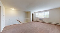This single-floor unit offers a practical and straightforward layout across several distinct rooms, each finished with neutral tones and functional details. The main living area is generously proportioned, featuring wall-to-wall carpet in a warm beige hue, a staircase with a white railing, and a large multi-pane window that allows natural light to fill the space. A wall-mounted air conditioning unit and baseboard heating ensure year-round climate control.

The kitchen is equipped with white cabinetry, dark laminate countertops in a granite-style finish, a built-in stovetop, a stainless steel sink, and an overhead range hood. The flooring transitions to a tile pattern, and an adjacent dining or eating area features a ceiling fan and open sightlines into the kitchen.

The bedrooms are carpeted and bright, with windows fitted with blinds. One bedroom includes a double sliding-door closet, while another offers direct access to a hallway. The bathrooms feature wood-look vinyl flooring, white subway tile wainscoting, and a combination tub and shower in the full bath, with a separate water closet visible in a secondary bathroom. Baseboard heating is consistent throughout the unit.