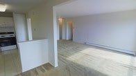 This single-floor residential space offers a clean, functional layout designed for comfortable everyday living. The open living area is finished with wall-to-wall neutral-toned carpeting that adds warmth underfoot and visually connects the space. Crisp white walls and a simple, understated color palette create a bright and adaptable interior ready for personalization.

A defining architectural feature is the partial wall column that partially divides the living area, lending a sense of structure while maintaining an open feel between spaces. The layout allows for a clear sightline from the main living zone through to a hallway corridor, which leads to additional rooms beyond. A pass-through counter is visible along one edge, suggesting a connection to a kitchen or utility area nearby.

The space is equipped with baseboard heating running along the perimeter walls, providing efficient climate control throughout the colder months. Interior lighting is provided through ceiling-mounted fixtures, maintaining a consistent and even illumination across the rooms.

Overall, the space presents a straightforward, move-in-ready environment with a neutral aesthetic that suits a wide range of furnishing styles. The single-level layout ensures ease of movement and accessibility throughout the entirety of the unit.