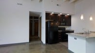 This single-floor loft showcases an industrial-modern aesthetic defined by dramatic soaring ceilings with exposed wooden beams and pristine white walls that amplify the sense of openness throughout the space. Polished concrete floors run seamlessly across the entire unit, contributing to the loft's contemporary, warehouse-inspired character.

The open-concept kitchen is a standout feature, equipped with rich dark espresso cabinetry, black appliances including a full-size refrigerator, electric range, and over-the-range microwave. A granite-topped breakfast bar separates the kitchen from the main living area, providing both casual dining space and a functional workspace. Recessed lighting and a pendant light above the bar illuminate the kitchen zone effectively.

The expansive main living area benefits from the soaring ceiling height, exposed structural beams, and an exposed brick accent wall that adds texture and a nod to the building's industrial origins. The layout flows naturally from the living space into a hallway leading to additional rooms, which feature warm wood-toned doors. The overall ambiance is open, airy, and distinctly urban, blending raw architectural elements with modern finishes to create a striking and functional living environment.