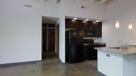 This single-floor loft showcases an industrial-modern aesthetic defined by dramatic soaring ceilings with exposed wooden beams and pristine white walls that amplify the sense of openness throughout the space. Polished concrete floors run seamlessly across the entire unit, contributing to the loft's contemporary, warehouse-inspired character.

The open-concept kitchen is a standout feature, equipped with rich dark espresso cabinetry, black appliances including a full-size refrigerator, electric range, and over-the-range microwave. A granite-topped breakfast bar separates the kitchen from the main living area, providing both casual dining space and a functional workspace. Recessed lighting and a pendant light above the bar illuminate the kitchen zone effectively.

The expansive main living area benefits from the soaring ceiling height, exposed structural beams, and an exposed brick accent wall that adds texture and a nod to the building's industrial origins. The layout flows naturally from the living space into a hallway leading to additional rooms, which feature warm wood-toned doors. The overall ambiance is open, airy, and distinctly urban, blending raw architectural elements with modern finishes to create a striking and functional living environment.