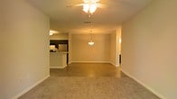 This single-floor space showcases an open-concept layout that seamlessly connects the living, dining, and kitchen areas into one cohesive living environment. The living area features soft, wall-to-wall carpeting underfoot, while the transition toward the dining and kitchen zones shifts to hard flooring, providing a natural visual boundary between spaces. A ceiling fan with integrated lighting fixture is centrally positioned above the living area, ensuring both comfort and illumination throughout. The dining area is defined by a hanging pendant light, adding a focused and functional overhead light source. The kitchen is positioned to the left and features dark cabinetry, a stainless steel refrigerator, and a raised breakfast bar that serves as both a workspace and casual dining surface. The kitchen's dark cabinet contrast against the lighter walls creates a defined, intentional design element. The entire space is painted in warm, neutral tones that complement the overall ambiance, giving the interior a welcoming and cohesive feel. A hallway visible to the right suggests access to additional rooms, completing this practical and efficiently designed single-level layout.