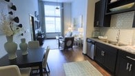 This single-floor space showcases a thoughtfully designed open-concept layout that seamlessly integrates the kitchen, dining, and living areas into one cohesive environment. Rich hardwood flooring runs throughout, providing a warm and grounding foundation for the entire space.

The kitchen features dark espresso cabinetry with modern brushed-nickel hardware, granite countertops, a tiled backsplash, and stainless steel appliances, including a dishwasher. The design is both functional and visually striking, with ample storage and clean lines.

Adjacent to the kitchen, the dining area comfortably accommodates a full table and chairs, making it suitable for everyday meals or entertaining. The transition into the living area is fluid, where tall windows with horizontal blinds and elegant draping allow abundant natural light to fill the room, creating an airy, open atmosphere.

The living area is anchored by tasteful furnishings and soft lighting, contributing to a comfortable and inviting ambiance. The generous ceiling height enhances the sense of spaciousness throughout. Overall, the space balances contemporary design with a warm, livable aesthetic.