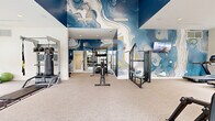 This fitness center in Nashville, TN offers a thoughtfully designed, single-floor workout facility that combines striking aesthetics with comprehensive equipment. The space is immediately distinguished by its dramatic, large-scale abstract murals rendered in deep navy blue, white, and gold tones, which adorn the walls and lend an artistic, energizing atmosphere to the environment. Soaring ceilings in the main workout area amplify the sense of openness, while recessed lighting ensures the space is well-illuminated throughout.

The facility is organized to support a full range of fitness activities. A dedicated strength-training zone features cable machines, a multi-function rack, a weight bench, a punching bag, TRX suspension trainers, and a dumbbell rack. A separate cardio area is lined with large windows that allow ample natural light and outdoor views of the surrounding neighborhood, and is equipped with treadmills, ellipticals, stair climbers, and stationary bikes. Wall-mounted televisions are positioned for convenient viewing during workouts.

The flooring throughout is soft, neutral-toned carpet, contributing to a clean and polished finish. A set of glass double doors provides access to an outdoor area, further enhancing the light-filled, open feel of the space. The overall design strikes a balance between functional utility and contemporary visual appeal.