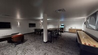 This single-floor entertainment and lounge space is designed to accommodate both social gatherings and casual recreation. The room features a neutral color palette of white walls and dark baseboards, complemented by textured, patterned carpet flooring that adds visual depth throughout. Recessed lighting and sleek wall-mounted sconces provide layered illumination, creating a versatile ambiance adaptable to various activities.

The space is anchored by multiple entertainment zones. A foosball table and a shuffleboard table serve as central recreational features, while a standalone arcade cabinet adds a retro gaming element. Multiple flat-screen televisions are wall-mounted throughout, ensuring clear sightlines from various seating positions. Seating options are varied and thoughtfully arranged, including a long communal high-top table with bar-height chairs for group gatherings, as well as smaller clusters of dark wood tables paired with modern orange upholstered chairs and lounge-style seating for more intimate settings.

A small kitchenette area with wood-toned cabinetry and a sink is tucked along one wall, providing functional convenience for guests. A structural column is integrated into the layout without interrupting the overall flow of the space. Bold geometric wall art adds a contemporary decorative touch, reinforcing the modern aesthetic throughout.