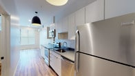 This single-floor unit showcases a thoughtfully designed layout with a consistent modern aesthetic carried throughout. The galley-style kitchen is a standout feature, outfitted with a full suite of stainless steel appliances including a refrigerator, electric range, built-in microwave, and dishwasher. Flat-front white cabinetry extends to the ceiling, maximizing storage, while dark stone countertops provide a striking contrast. A bold black dome pendant light adds a contemporary accent overhead. Wood-look flooring runs continuously throughout the unit, lending warmth and visual cohesion to each space. The open living area benefits from a wide, multi-panel window fitted with horizontal blinds, allowing generous natural light to fill the room. A wall-mounted mini-split HVAC unit provides efficient climate control. A separate room features a horizontal clerestory window, bringing in additional light while maintaining privacy. Recessed lighting is present throughout, ensuring well-lit interiors. The bathroom includes a full-size tub and shower combination with clean white surround and chrome fixtures. Neutral wall tones and clean architectural lines contribute to the unit's bright, minimalist ambiance.
