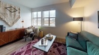 This single-floor unit offers a thoughtfully designed layout that balances comfort and modern style throughout. The living area is anchored by large windows that frame urban views and invite generous natural light, creating a warm and inviting atmosphere. Wood-look flooring runs seamlessly through the space, complementing the clean, neutral palette of the walls.

The kitchen is a standout feature, showcasing rich dark espresso cabinetry paired with light countertops and a classic tile backsplash. Stainless steel appliances — including a refrigerator, range, microwave, and dishwasher — provide a sleek, contemporary finish. A freestanding kitchen island with bar seating adds both additional prep space and a casual dining option.

The bathroom is well-proportioned and features matching dark wood cabinetry with a single-basin vanity, wood-look flooring, and a shower-tub combination. A teal accent wall adds a touch of color and character. Notably, a stacked in-unit washer and dryer is conveniently located just off the bathroom, adding everyday practicality.

The dollhouse view reveals a compact but efficiently arranged floor plan that makes excellent use of every area. Overall, the space presents a clean, modern aesthetic with functional amenities well-suited for comfortable everyday living.