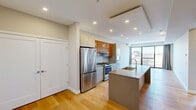 This well-appointed single-floor residence offers a thoughtfully designed open-concept layout that maximizes its efficient footprint. The space includes a living room, kitchen, one bedroom, one bathroom, a closet, and two hallways that connect the layout seamlessly.

The kitchen is a standout feature, showcasing a large quartz-topped island with an integrated sink, rich wood-grain upper cabinetry contrasted by sleek light gray lower cabinets, a subway tile backsplash, and a full suite of stainless steel appliances including a gas range, refrigerator, and built-in microwave. The kitchen flows directly into the living area, creating an open and connected common space.

The living room is bright and airy, benefiting from floor-to-ceiling sliding glass doors that open to a private balcony, offering open urban views and abundant natural light. Light hardwood flooring runs throughout the entire unit, contributing to a warm, cohesive aesthetic.

The bedroom is generously sized, also featuring large sliding glass doors with balcony access and urban views, along with a dedicated closet. The bathroom is finished with large-format linear tile, a glass-enclosed walk-in shower with a pebble floor, and contemporary fixtures throughout. Recessed lighting is present in every room, maintaining a clean, modern ambiance throughout the residence.