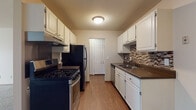 This single-floor unit offers a practical and comfortable layout with thoughtfully designed spaces throughout. The galley-style kitchen is a standout feature, lined on both sides with crisp white cabinetry offering generous storage. Dark countertops provide a striking contrast, while a mosaic tile backsplash in warm earth tones adds a decorative touch. The kitchen is equipped with a gas range, black refrigerator, and dishwasher, all set atop warm wood-look flooring. A closet door at the end of the kitchen adds convenient additional storage. The open living area is carpeted and bathed in a neutral palette, creating a calm and versatile space. A baseboard heater runs along the wall, and the room offers ample space to arrange furnishings comfortably. The bedroom continues the soft carpeted aesthetic and is fitted with a ceiling fan for year-round comfort. Two doors open to a walk-in closet and to the main hallway, providing excellent organization. The bathroom features tile flooring, a granite-look vanity countertop with white cabinetry, and a full tub-shower combination with white subway tile surround. Throughout the unit, consistent warm-toned walls and wood-accented trim tie the spaces together cohesively.