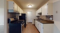 This single-floor unit offers a practical and comfortable layout with thoughtfully designed spaces throughout. The galley-style kitchen is a standout feature, lined on both sides with crisp white cabinetry offering generous storage. Dark countertops provide a striking contrast, while a mosaic tile backsplash in warm earth tones adds a decorative touch. The kitchen is equipped with a gas range, black refrigerator, and dishwasher, all set atop warm wood-look flooring. A closet door at the end of the kitchen adds convenient additional storage. The open living area is carpeted and bathed in a neutral palette, creating a calm and versatile space. A baseboard heater runs along the wall, and the room offers ample space to arrange furnishings comfortably. The bedroom continues the soft carpeted aesthetic and is fitted with a ceiling fan for year-round comfort. Two doors open to a walk-in closet and to the main hallway, providing excellent organization. The bathroom features tile flooring, a granite-look vanity countertop with white cabinetry, and a full tub-shower combination with white subway tile surround. Throughout the unit, consistent warm-toned walls and wood-accented trim tie the spaces together cohesively.