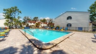 This outdoor pool area in Jacksonville, Florida offers a well-appointed recreational space designed for relaxation and leisure. The centerpiece is a generously sized rectangular swimming pool with clear blue water, accessible via stainless steel entry rails and surrounded by a broad, textured paver deck that provides ample room for movement and sunbathing.

The deck is furnished with multiple blue lounge chairs and chaise lounges positioned along the poolside, along with a set of colorful chairs and a table clustered near a handsome wood-toned pergola. The pergola serves as a shaded gathering point, adding architectural interest to the open-air setting. Additional seating areas are distributed around the perimeter, accommodating a variety of uses throughout the day.

Mature trees and tropical plantings, including sago palms and ornamental shrubs set in mulched beds, frame the space and contribute a lush, Florida-appropriate aesthetic. The area is enclosed by a black wrought iron fence with gated entry, ensuring both security and visual definition. The surrounding residential buildings are visible beyond the fence, providing a sense of community context. Overall, the space offers a bright, open, and inviting outdoor environment well-suited to the warm Jacksonville climate.