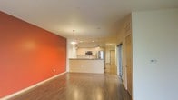 This single-floor space is designed with an open-concept layout that seamlessly connects the living area, dining space, and kitchen into one cohesive environment. A striking terracotta-orange accent wall anchors the living area, adding warmth and a bold pop of color that contrasts beautifully against the crisp white ceilings and neutral-toned walls throughout the rest of the space.

Rich, dark hardwood flooring runs continuously across the entire floor, lending a sense of warmth and continuity. The kitchen features a peninsula-style counter with a dark countertop, light wood cabinetry, stainless steel appliances including a refrigerator and microwave, and a sink with clean sightlines into the main living area. Pendant lighting hangs elegantly above the kitchen peninsula, complementing the recessed lighting that illuminates the space overhead.

The layout is notably spacious and airy, with generous room to move between areas. Large sliding glass doors are visible toward the back of the space, offering natural light and potential access to an outdoor area. Additional doors suggest private rooms or closet spaces beyond the open living zone. Overall, the space strikes a balance between modern design and a welcoming, livable atmosphere.