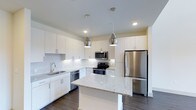 This two-story Dallas property showcases a thoughtfully designed interior with contemporary finishes throughout. The kitchen serves as a standout feature, boasting crisp white cabinetry that extends to the ceiling for ample storage, complemented by a textured white tile backsplash that adds subtle visual interest. A granite-topped central island provides additional prep space and casual seating potential, while matching granite countertops run along the perimeter. Stainless steel appliances, including a French door refrigerator, electric range, built-in microwave, and dishwasher, lend a sleek, professional feel. Two polished metallic pendant lights hang above the island, enhancing both function and style. Recessed lighting illuminates the space evenly, and dark hardwood flooring creates a striking contrast against the white palette. The kitchen's open layout connects seamlessly to adjacent living areas, contributing to an airy, modern atmosphere. Spanning two floors, the overall layout offers versatile living arrangements suited for both everyday comfort and entertaining.