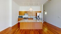 This ADA-compliant one-bedroom, one-bathroom residence offers a thoughtfully designed, single-floor layout with a strong emphasis on accessibility and modern finishes throughout. Rich hardwood flooring flows seamlessly across the living, kitchen, and bedroom areas, lending warmth and continuity to the open-concept floor plan.

The kitchen is a standout feature, equipped with warm wood cabinetry, granite countertops, a large central island with pendant lighting overhead, stainless steel appliances including a side-by-side refrigerator with ice maker, a range, dishwasher, and microwave. Track lighting illuminates the space efficiently, while a charming arched doorway adds an architectural detail along the hallway leading to the private areas.

The living and dining area benefits from generous floor-to-ceiling windows that frame vibrant urban views, filling the space with abundant natural light. The bedroom continues the hardwood flooring and features large windows with striking city views, creating an airy and inviting atmosphere.

The bathroom is designed with accessibility as a priority, featuring a roll-in shower with stainless steel grab bars, a handheld showerhead, built-in corner shelving, and neutral tile surround. A granite-topped vanity with wood cabinetry and a large mirror completes the space. An in-unit washer is also present, adding everyday convenience.