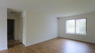 This single-floor space presents a clean, minimalist aesthetic designed to maximize comfort and functionality. The main living area is characterized by smooth white walls and warm hardwood flooring that runs throughout, lending a sense of continuity and warmth to the interior. Natural light flows in through a large window fitted with vertical blinds, allowing for adjustable light control while offering views of the surrounding greenery outdoors. The ceiling is well-proportioned, contributing to the airy, open feel of the room. A door with visible trim detail leads to an adjacent space, suggesting a practical layout with defined zones. The overall ambiance is bright and neutral, providing a versatile canvas well-suited to a variety of interior styles and furnishing choices. The crisp white surfaces and natural wood tones combine to create a welcoming atmosphere that feels both fresh and timeless. Electrical outlets are thoughtfully positioned along the baseboards, and a ceiling vent ensures proper climate control throughout the space. This well-maintained interior offers a straightforward, functional design with a calm and inviting character.