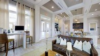 This single-floor space showcases a refined interior design that blends coastal and contemporary aesthetics. The most striking architectural feature is the coffered ceiling, finished with wood-planked insets in a warm gray tone, contrasted by crisp white trim and recessed lighting that illuminates the space evenly throughout.

The open layout connects multiple functional zones seamlessly. A comfortable lounge area anchors the main space, furnished with a rich brown leather sofa accented by soft blue and white throw pillows. Decorative candles and organic branch arrangements add an understated elegance to the setting. Nearby, a dining or gathering area with a light-toned table adds versatility.

A dedicated workstation nook near the perimeter features a built-in desk with bar-height seating, ideal for focused tasks. Light-washed hardwood flooring runs throughout, enhancing the airy, bright atmosphere.

Glass-paned French doors draw in natural light and provide views to an exterior area, contributing to a sense of openness. Sheer linen drapery softens the windows, while pendant lighting adds warmth to adjacent areas. Overall, the space exudes a calm, welcoming ambiance with thoughtful design details throughout.