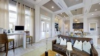This single-floor space showcases a refined interior design that blends coastal and contemporary aesthetics. The most striking architectural feature is the coffered ceiling, finished with wood-planked insets in a warm gray tone, contrasted by crisp white trim and recessed lighting that illuminates the space evenly throughout.

The open layout connects multiple functional zones seamlessly. A comfortable lounge area anchors the main space, furnished with a rich brown leather sofa accented by soft blue and white throw pillows. Decorative candles and organic branch arrangements add an understated elegance to the setting. Nearby, a dining or gathering area with a light-toned table adds versatility.

A dedicated workstation nook near the perimeter features a built-in desk with bar-height seating, ideal for focused tasks. Light-washed hardwood flooring runs throughout, enhancing the airy, bright atmosphere.

Glass-paned French doors draw in natural light and provide views to an exterior area, contributing to a sense of openness. Sheer linen drapery softens the windows, while pendant lighting adds warmth to adjacent areas. Overall, the space exudes a calm, welcoming ambiance with thoughtful design details throughout.