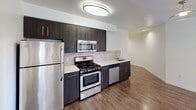 This contemporary single-floor residence offers a well-designed open-concept layout that seamlessly connects the kitchen, dining, and living areas. Rich hardwood-style flooring runs throughout the main living spaces, contributing to a warm and cohesive aesthetic. The kitchen is a standout feature, outfitted with dark espresso cabinetry, a textured tile backsplash, light countertops, and a full suite of stainless steel appliances including a gas range, microwave, refrigerator, and dishwasher. A peninsula with a stainless steel sink provides additional prep space and a natural boundary between the kitchen and living area. The living room benefits from abundant natural light, with French doors opening to an outdoor area framed by lush greenery. The bedroom is finished with plush carpeting, a ceiling fan, and a large window with views of the surrounding landscape. Two bathrooms serve the residence — one featuring a soaking tub with tiled surround accented by a decorative mosaic border and an LED-lit vanity mirror, while the second offers a clean, all-white design with tile flooring. A dedicated in-unit laundry closet with a stacked washer and dryer adds everyday convenience. The overall ambiance is clean, modern, and thoughtfully appointed.