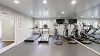 Located in Lorton, VA, this dedicated fitness center offers a well-equipped exercise environment designed to support a range of workout routines. The space is divided into distinct functional zones, with a main cardio area housing multiple treadmills and elliptical machines by True and other brands, alongside a separate strength training section outfitted with a cable machine system, a flat workout bench, free weights, and a yoga mat. Floor-to-ceiling mirrors line the walls, visually expanding the room and allowing users to monitor their form during exercise. The durable rubber flooring throughout provides both safety and comfort underfoot. Recessed lighting illuminates the entire space evenly, and a flat-screen television is mounted on the wall for entertainment during workouts. A water dispenser station is conveniently positioned near the entry door, which features glass sidelights that allow natural light to filter in. Windows along one wall further enhance the brightness of the room. A decorative column serves as an architectural accent while subtly delineating the cardio and strength zones. The clean, neutral color palette of white and gray walls contributes to a focused and functional atmosphere.