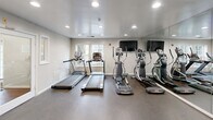 Located at 9454 Orange Blossom Trail in Lorton, Virginia, this fitness center is a thoughtfully organized exercise facility designed to support a full range of workout routines. The space is laid out across a single floor and is divided into distinct functional zones, maximizing utility within a compact footprint.

The main cardio area features a row of elliptical trainers and treadmills — including True-brand treadmills — arranged neatly along one side of the room. Floor-to-ceiling mirrors line the walls, amplifying the sense of space and allowing users to monitor their form during exercise. Recessed ceiling lighting provides bright, even illumination throughout, while a wall-mounted flat-screen television adds an entertainment option for cardio sessions.

A separate strength training alcove houses a multi-station cable machine, a flat workout bench, a yoga mat, and a small weight rack, offering a dedicated area for resistance training. The space benefits from natural light via windows along one wall, and a water dispenser station is conveniently positioned near the entrance for easy access.

Underfoot, durable rubber flooring covers the entire exercise area, providing both safety and comfort. Decorative white columns add a refined architectural touch to the otherwise clean, neutral interior. An adjacent door leads to a secondary room, further extending the facility's utility. Overall, the fitness center presents a clean, functional, and well-maintained environment suited for daily workouts.