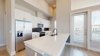 This thoughtfully designed 2-bedroom, 2-bathroom unit offers a modern aesthetic with clean lines and contemporary finishes throughout. The open-concept kitchen is a standout feature, boasting white cabinetry with matte black hardware, quartz countertops, a large peninsula with an integrated sink, stainless steel appliances, and a dark tile backsplash. A built-in open pantry provides generous additional storage. Light wood-style flooring flows seamlessly through the entire unit, complementing the neutral warm-toned walls.

The living area impresses with soaring double-height ceilings and floor-to-ceiling windows on two walls, flooding the space with natural light and offering open exterior views. Both bedrooms are well-proportioned, with the second bedroom featuring dual windows that bring in ample daylight.

The primary bathroom is elegantly appointed with a dual-sink white vanity, matte black fixtures, a framed mirror with lighted sconces, and a glass-enclosed tiled shower. The second bathroom features a soaking tub with full tile surround and a single-sink vanity. Both bathrooms provide direct access to dedicated closets, maximizing storage and convenience. A hallway entry nook with built-in hooks adds a practical finishing touch.