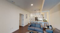This thoughtfully designed single-floor residence combines modern aesthetics with functional living. The open-concept layout seamlessly connects the living room and kitchen, creating an inviting, airy atmosphere enhanced by elevated ceilings and recessed lighting. The living area features rich dark hardwood flooring, a neutral color palette, and generous natural light, complemented by a stylish grid-frame mirror as a focal design element.

The kitchen is a standout feature, showcasing two-tone cabinetry with white upper cabinets and warm wood-toned lower cabinets, all accented by sleek matte black hardware. Stainless steel appliances, including a refrigerator, range, and microwave, are paired with crisp white quartz countertops and a classic subway tile backsplash. A peninsula with pendant lighting provides additional seating and workspace, flowing naturally into the dining and living areas.

The bedrooms are calm and comfortable, each featuring large windows that bring in natural light and offer views of the outdoor surroundings, including a private balcony accessible from one of the rooms. The bathroom is finished with wood-toned cabinetry, a well-lit vanity mirror, matte black fixtures, and durable wood-look flooring, achieving a clean, modern look throughout.