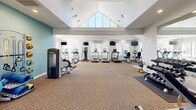 Located at 300 NC-146 in Asheville, NC, this single-floor fitness center is thoughtfully designed to accommodate a comprehensive range of workout activities. The facility is organized into distinct functional zones, each outfitted with purpose-appropriate equipment.

The main strength training area commands attention with its dramatic vaulted ceiling, recessed lighting, and a ceiling fan that promotes air circulation. Large triangular windows near the roofline flood the space with natural light. This zone houses cable resistance machines, benches, a dumbbell rack, barbells, stability balls, TRX suspension equipment, and kettle bells, all arranged across durable carpeted flooring.

A dedicated cardio zone features a row of treadmills, elliptical trainers, and stationary bikes by TRUE Fitness, positioned along windowed walls to maximize natural light. Wall-mounted televisions are positioned throughout for entertainment during workouts.

A separate entry area with tile flooring provides a clean transition point, complete with a hand sanitizer station and exterior door access. The overall aesthetic is clean and bright, with neutral gray and white walls accented by soft teal tones, creating a motivating and welcoming workout environment.