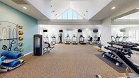 Located at 300 NC-146 in Asheville, NC, this single-floor fitness center is thoughtfully designed to accommodate a comprehensive range of workout activities. The facility is organized into distinct functional zones, each outfitted with purpose-appropriate equipment.

The main strength training area commands attention with its dramatic vaulted ceiling, recessed lighting, and a ceiling fan that promotes air circulation. Large triangular windows near the roofline flood the space with natural light. This zone houses cable resistance machines, benches, a dumbbell rack, barbells, stability balls, TRX suspension equipment, and kettle bells, all arranged across durable carpeted flooring.

A dedicated cardio zone features a row of treadmills, elliptical trainers, and stationary bikes by TRUE Fitness, positioned along windowed walls to maximize natural light. Wall-mounted televisions are positioned throughout for entertainment during workouts.

A separate entry area with tile flooring provides a clean transition point, complete with a hand sanitizer station and exterior door access. The overall aesthetic is clean and bright, with neutral gray and white walls accented by soft teal tones, creating a motivating and welcoming workout environment.