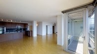 This single-floor space showcases a generous open-plan layout with light hardwood flooring that extends throughout, creating a warm, cohesive aesthetic. Structural columns punctuate the living area, adding architectural character while defining zones within the open floor plan. The kitchen, positioned to one side, features dark wood cabinetry, dark countertops, and a stainless steel sink, offering a contemporary contrast to the bright, neutral walls and light flooring. Recessed lighting in the ceiling contributes to a clean, uncluttered overhead profile. A standout feature is the floor-to-ceiling sliding glass door system on one wall, fitted with a slim aluminum frame, which opens to an outdoor space — likely a balcony or terrace — allowing natural light to filter into the interior. Doorways visible toward the rear of the space suggest additional rooms or utility areas beyond the main living zone. The overall ambiance is modern and minimalist, with an emphasis on open space, natural light, and a neutral palette that provides a versatile foundation for a variety of uses.
