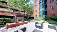 The outdoor courtyard at Symphony Center Apartments is a thoughtfully designed residents-only amenity space nestled within a classic urban brick building complex. The courtyard is framed by warm red brick architecture and tall glass-and-steel facades, creating an enclosed yet open-air environment that balances privacy with an urban atmosphere.

At the heart of the space is a prominent brick fireplace, serving as a central gathering focal point and providing a cozy ambiance. Surrounding it are multiple seating areas, including wicker lounge chairs with neutral cushions and green metal benches, accommodating both relaxed lounging and casual social gatherings.

Mature trees provide generous shade throughout the paved courtyard, while curated landscaping, including low shrubs, ground cover plantings, and potted greenery near the entrance, adds lush natural texture to the environment. The paver stone flooring runs throughout, maintaining a clean and cohesive aesthetic.

Access to the courtyard is through teal-framed glass doors marked "Residents Only," reinforcing the exclusive and secure nature of the space. The combination of natural greenery, architectural warmth, and well-chosen furnishings creates an inviting urban retreat ideal for outdoor relaxation.